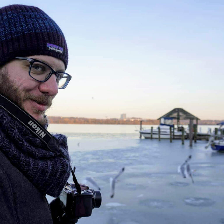 Joe is seen half-way turned towards the camera with his own camera in hand as he overlooks the frozen bay in Alexandria, VA.